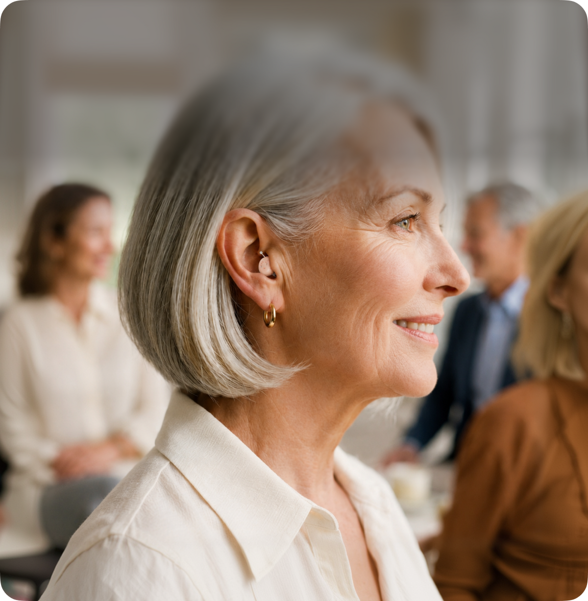 Woman with gray hair smiling in a social setting with hearing device in her ear 